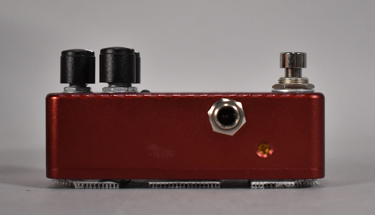 The red metallic pedal is viewed from the side, showing two black knobs, a chrome input jack, a small amber LED, and a footswitch.