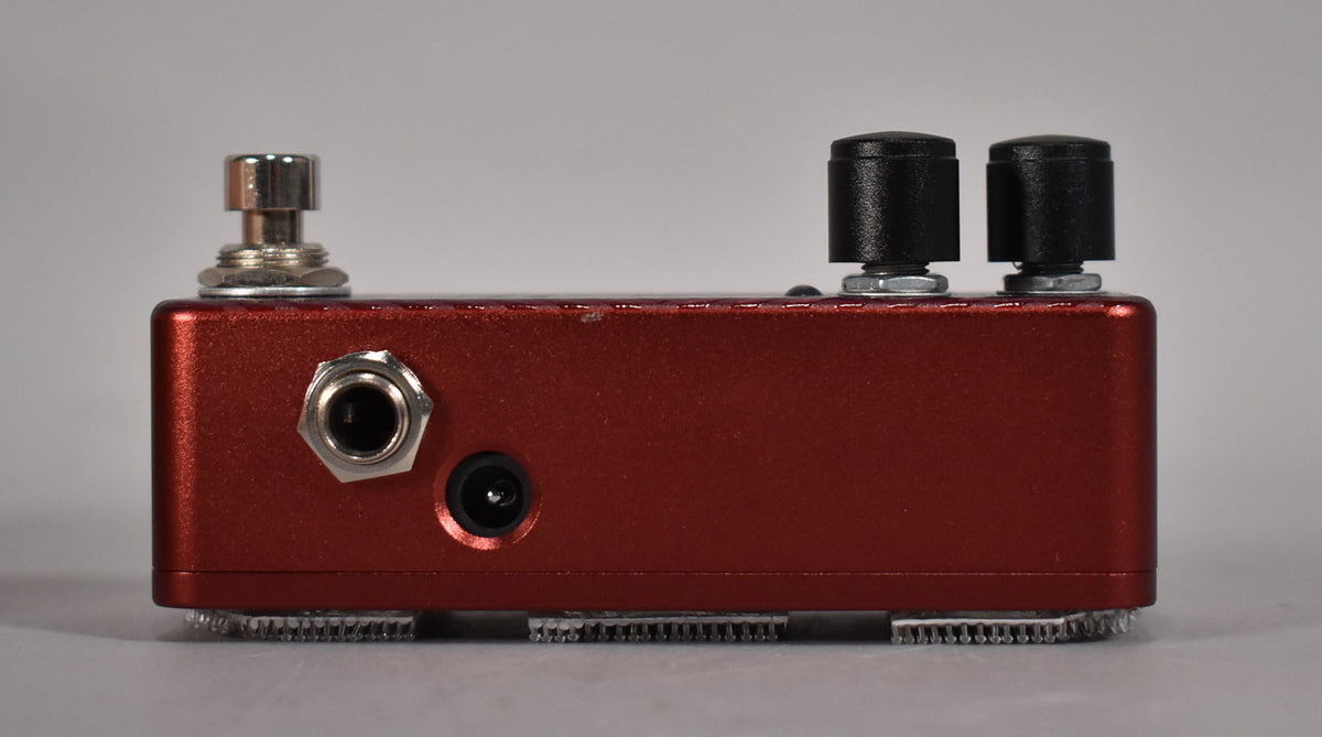 The cranberry red pedal is seen from the side, showing the input jack, power input, and two black control knobs on top.
