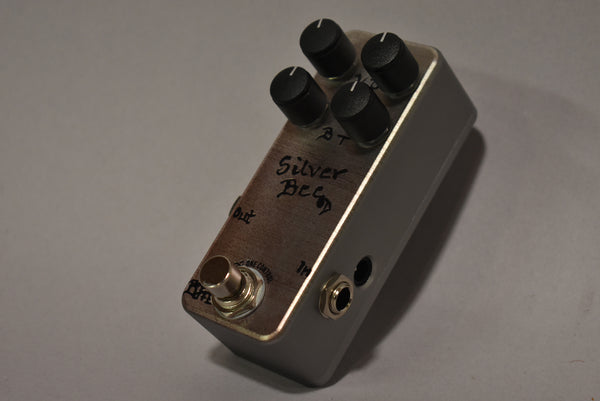 A silver guitar pedal, viewed from above and angled right, with four black control knobs and two input jacks.