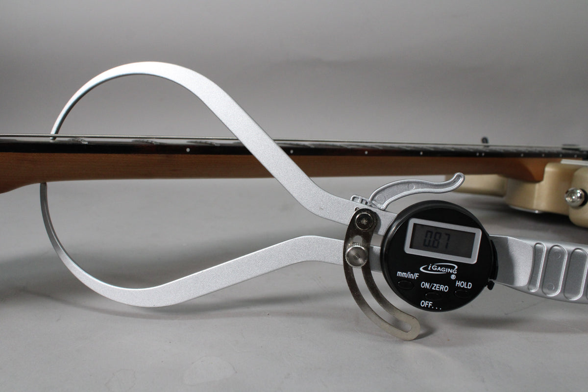 A digital caliper measures the guitar's neck, showing a reading of 0.87, with the fretboard and body visible in the background.