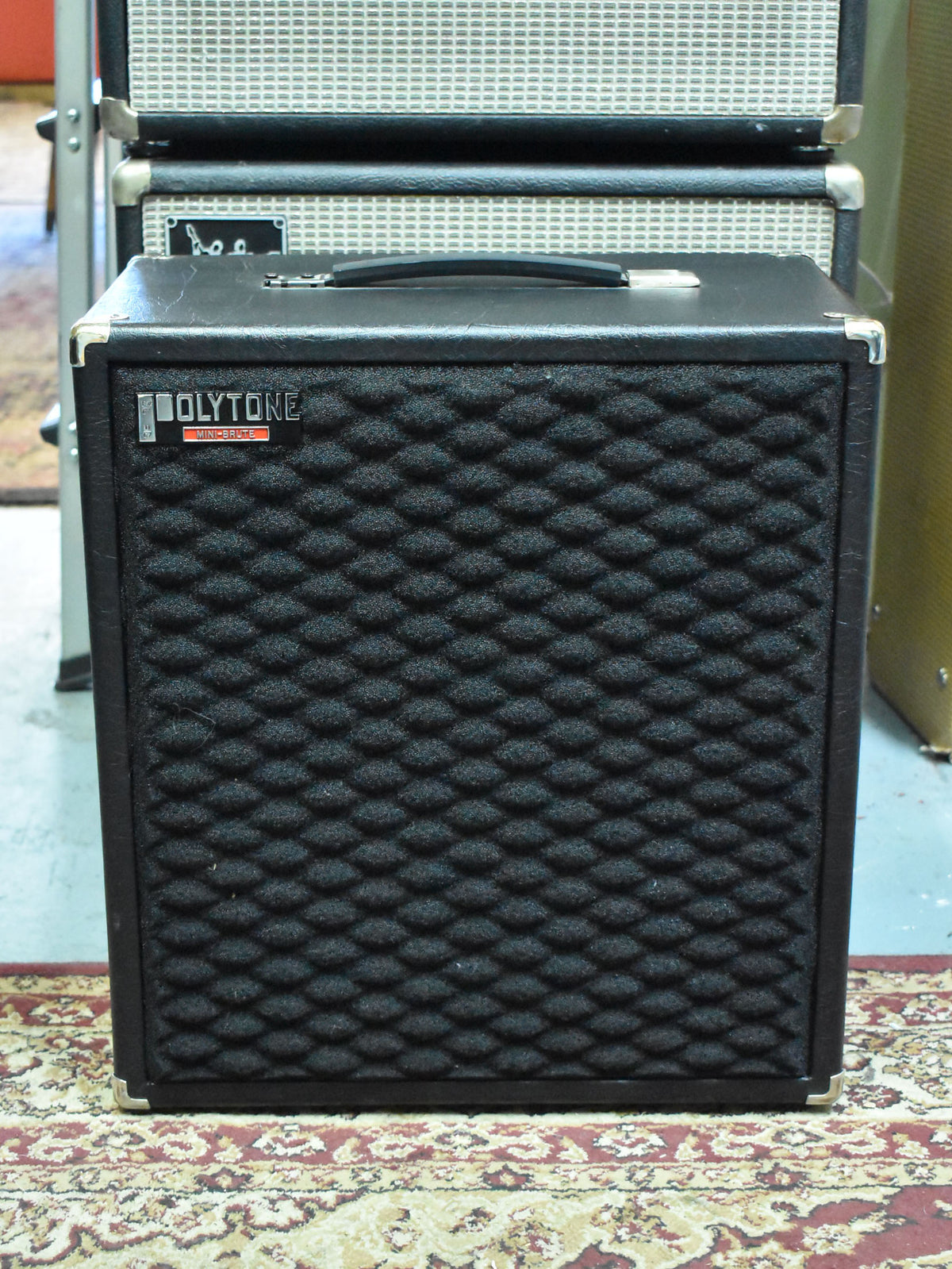 Front view of a black speaker cabinet with a textured, dimpled grille, chrome corner protectors, and a top handle.