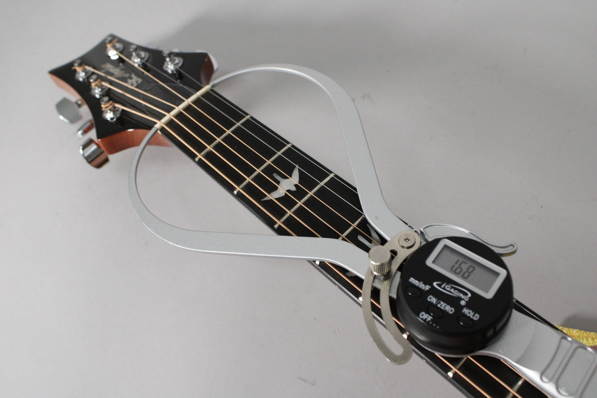 A digital caliper measures the string height on the dark rosewood fretboard, showing a bird inlay at the 12th fret.