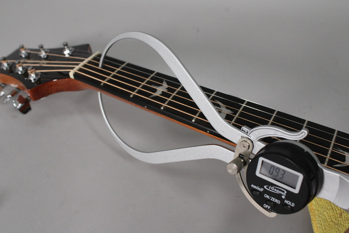 A digital caliper measures the neck relief of the guitar, showing 0.93mm, with bird inlays visible on the dark fretboard.