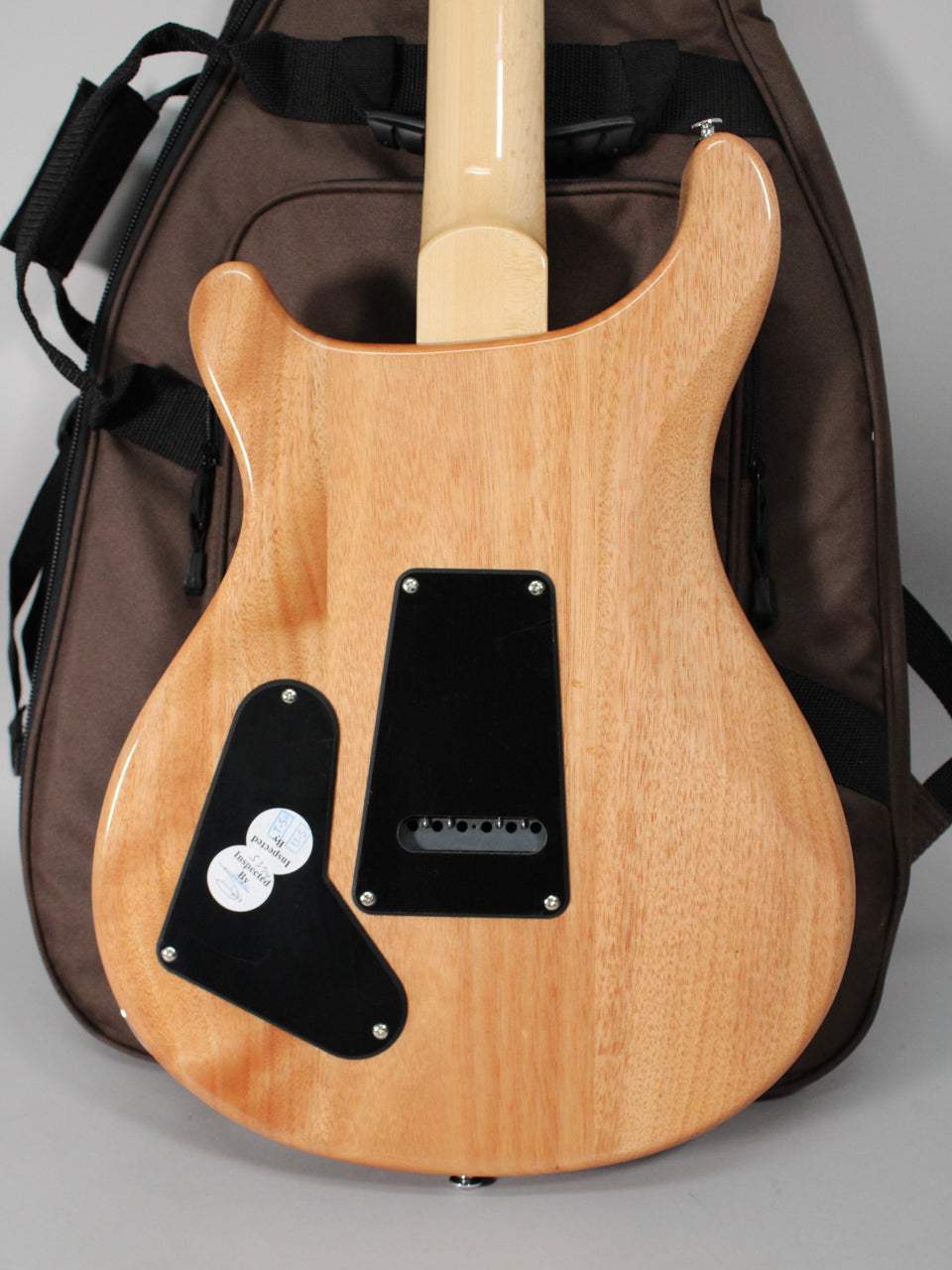 The guitar's light brown wooden back, with a glossy finish, shows two black control plates and a light maple neck, resting against a brown gig bag.