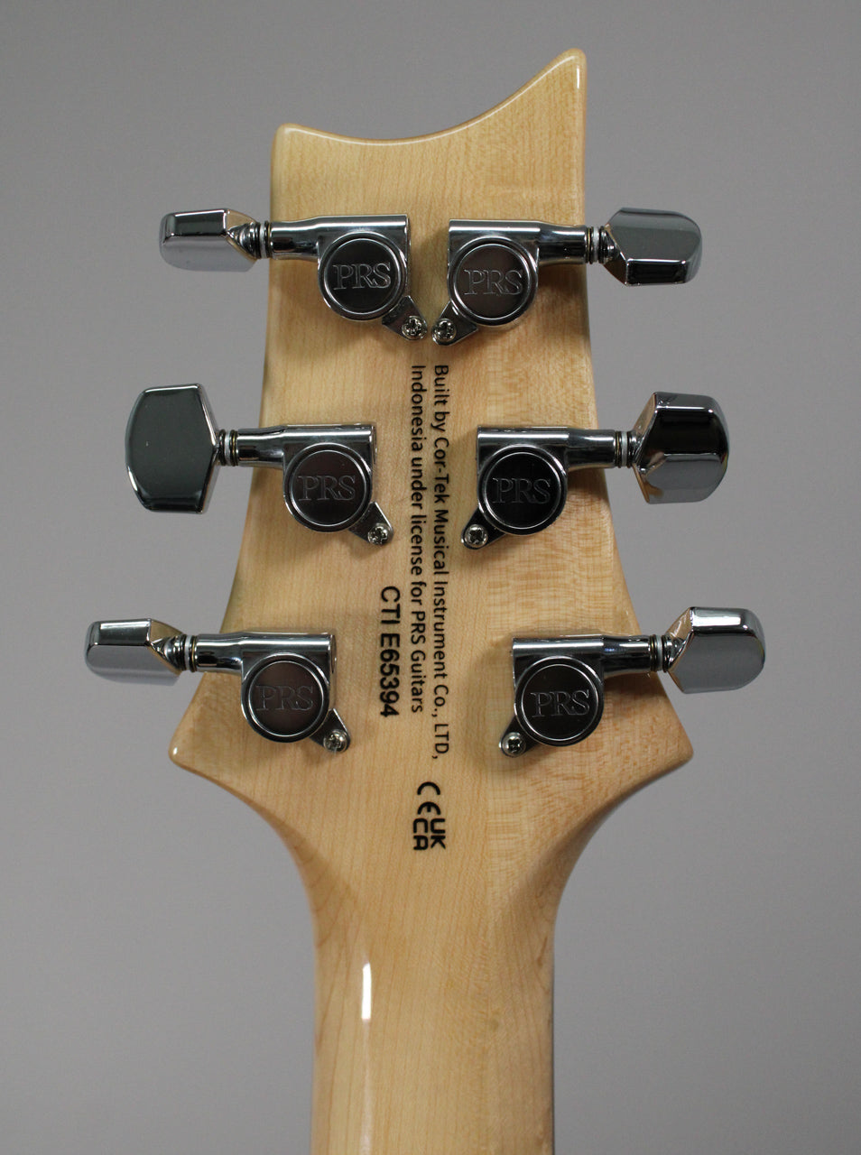 The back of a light wood guitar headstock with six chrome tuners, three on each side, and black text printed vertically.