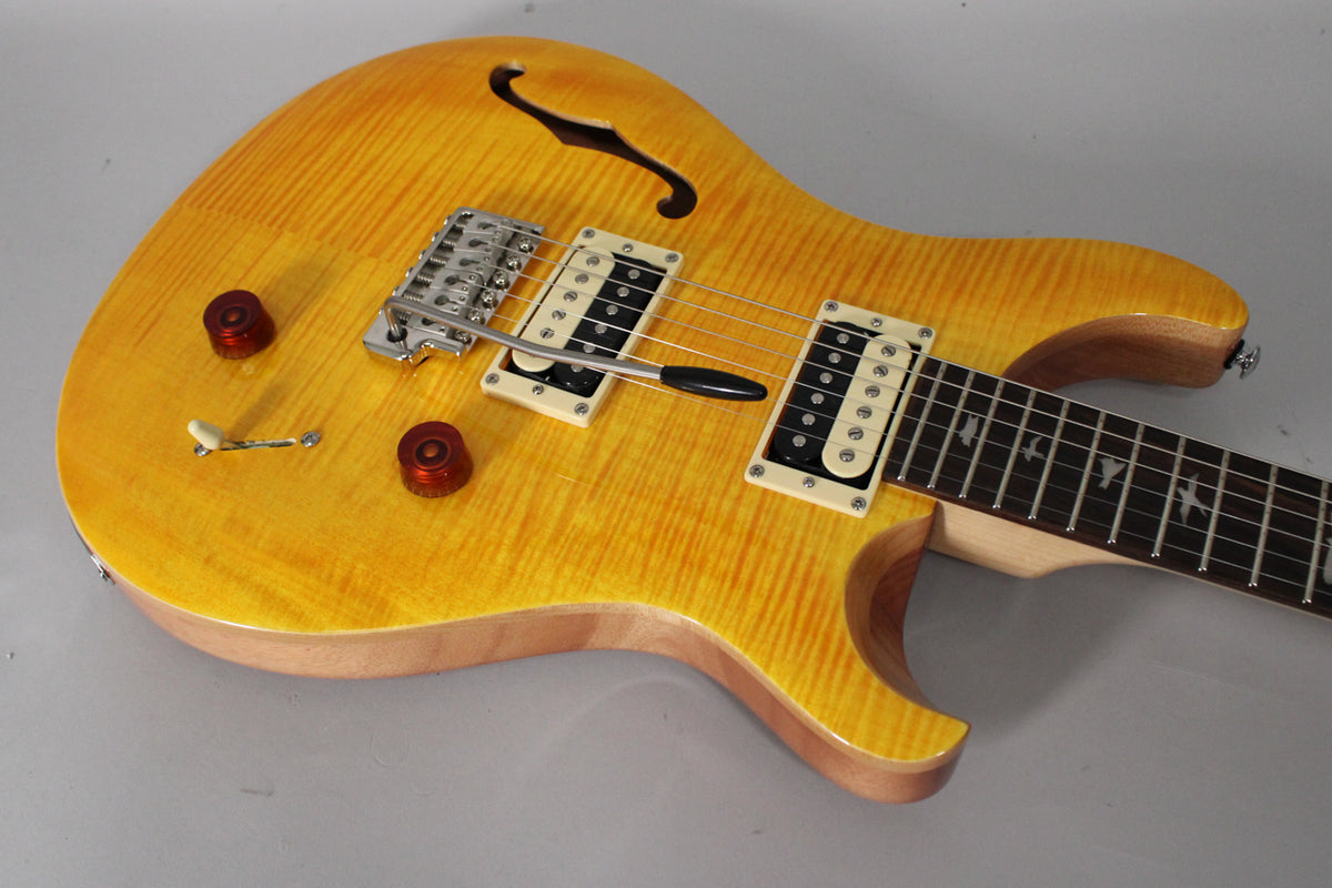 The Santana Yellow semi-hollow guitar, angled from the upper right, shows its f-hole, two humbuckers, tremolo, and two amber knobs.