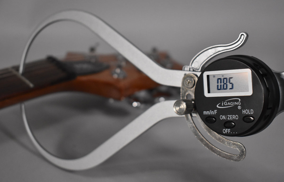 A digital caliper measures the guitar's fretboard, showing 0.85 on its screen, with the headstock blurred in the background.