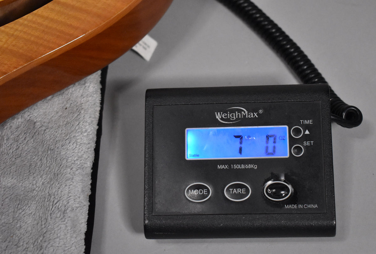 The guitar's lower bout, with a sunburst finish and visible wood grain, rests on a grey cloth next to a digital scale displaying "7 lb 0 oz".