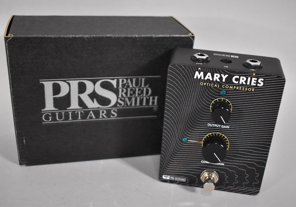 A black guitar pedal with two gold-ringed black knobs and a silver footswitch, featuring a wavy line pattern and a partially visible box.