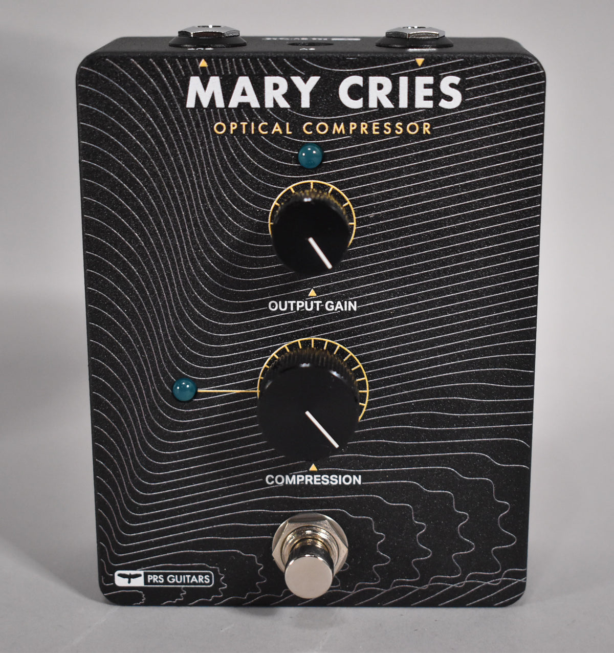 A black guitar pedal with white topographic lines, two black knobs with gold markings, and a silver footswitch.