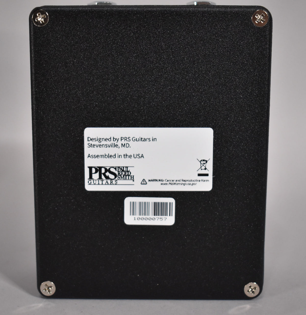 The black textured pedal is viewed from the back, showing a white label with text and a barcode, secured by four silver screws.