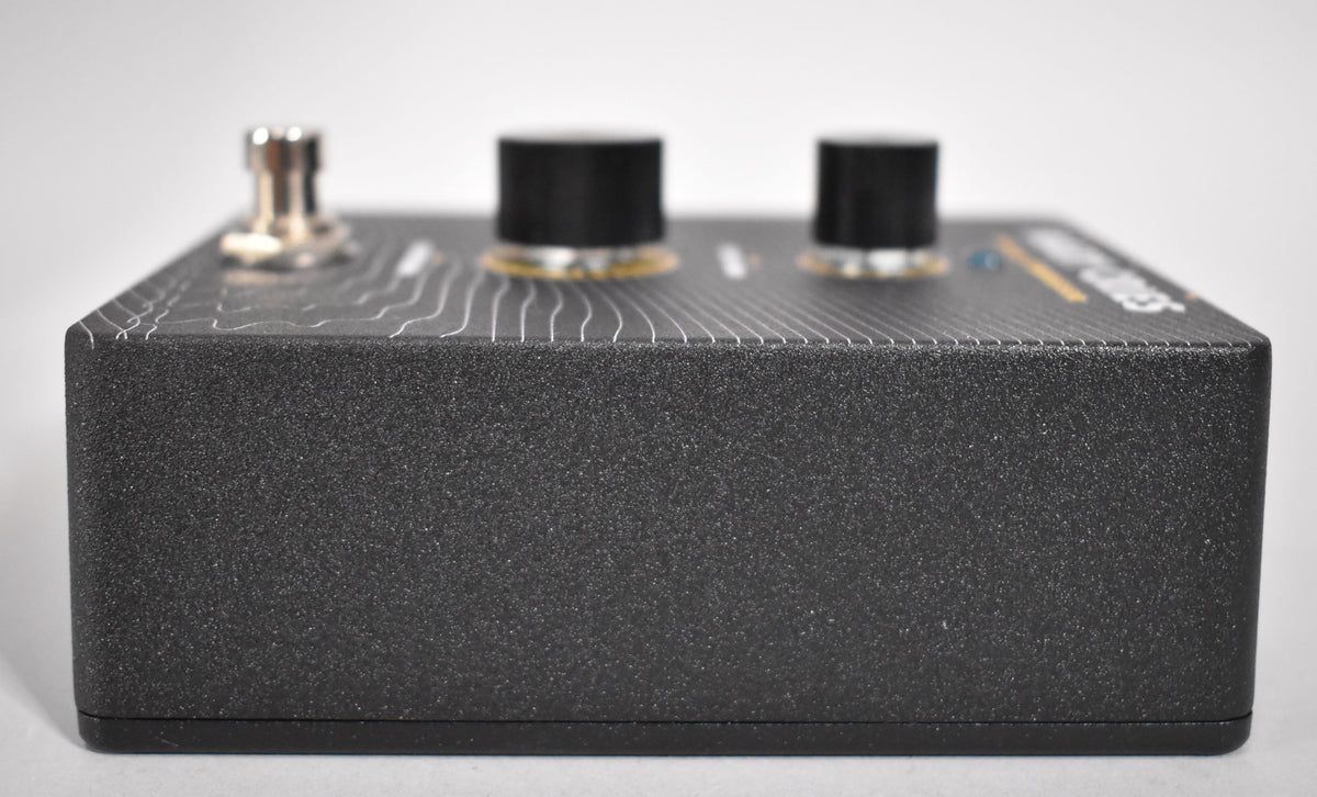 The black textured pedal is viewed from the right side, showing a silver footswitch and two black knobs on the top surface.