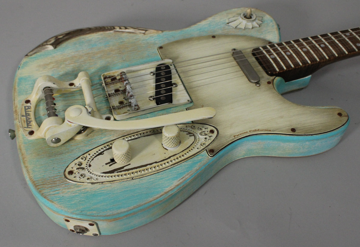 The guitar's body, angled from above, shows a heavily relic'd blue and cream finish, a Bigsby tremolo, and ornate control plate.
