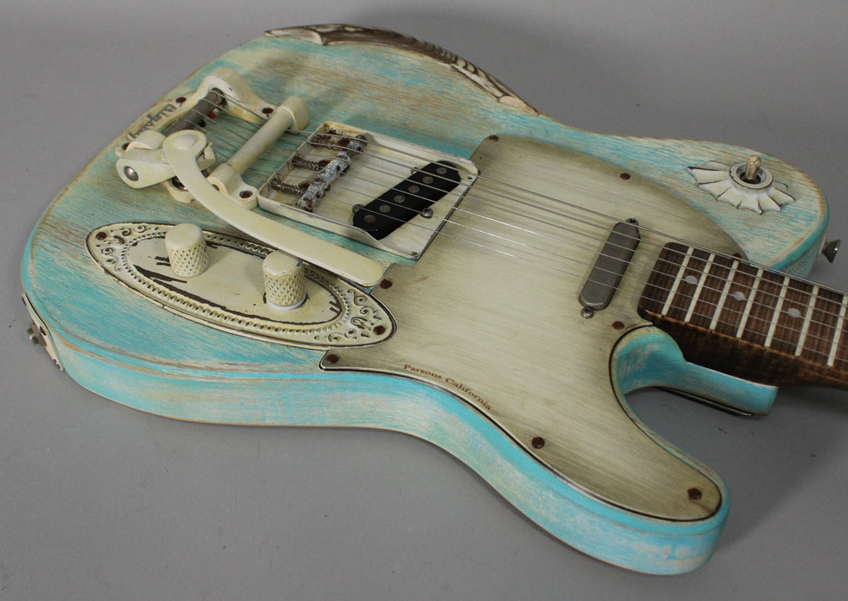 Close-up of a blue and cream relic guitar body with a Bigsby tremolo, two pickups, and two knurled knobs on an ornate plate.