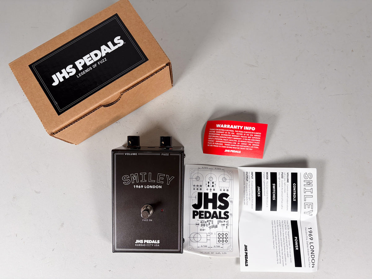 Overhead view of a dark gray fuzz pedal with two black knobs, a footswitch, and a red indicator light, next to its box and papers.