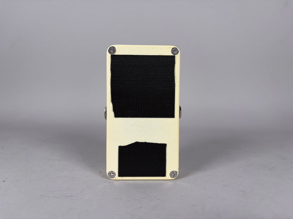 The cream-colored pedal is seen from the back, with black hook-and-loop strips covering most of the surface and four silver screws.