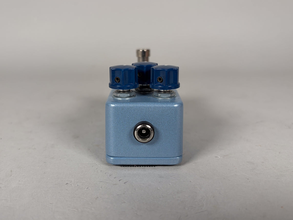The light blue metallic pedal is viewed from the back, showing a power input jack and three dark blue control knobs.