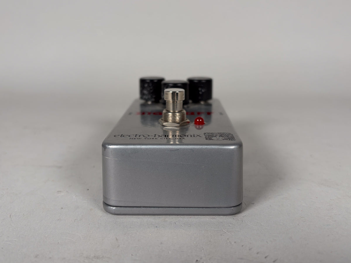 The silver pedal is viewed from the front, showing the footswitch, three black control knobs, and a small red LED light on the top surface.