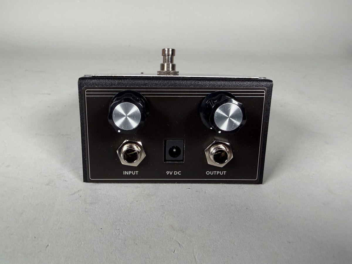 The black guitar pedal is viewed from the front, showing two silver-capped black knobs, input/output jacks, and a 9V DC power port.