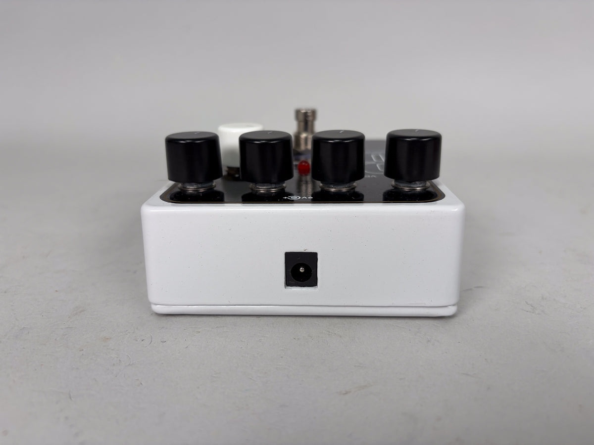 The white pedal is viewed from the front, showing the power input jack and four black control knobs on top.