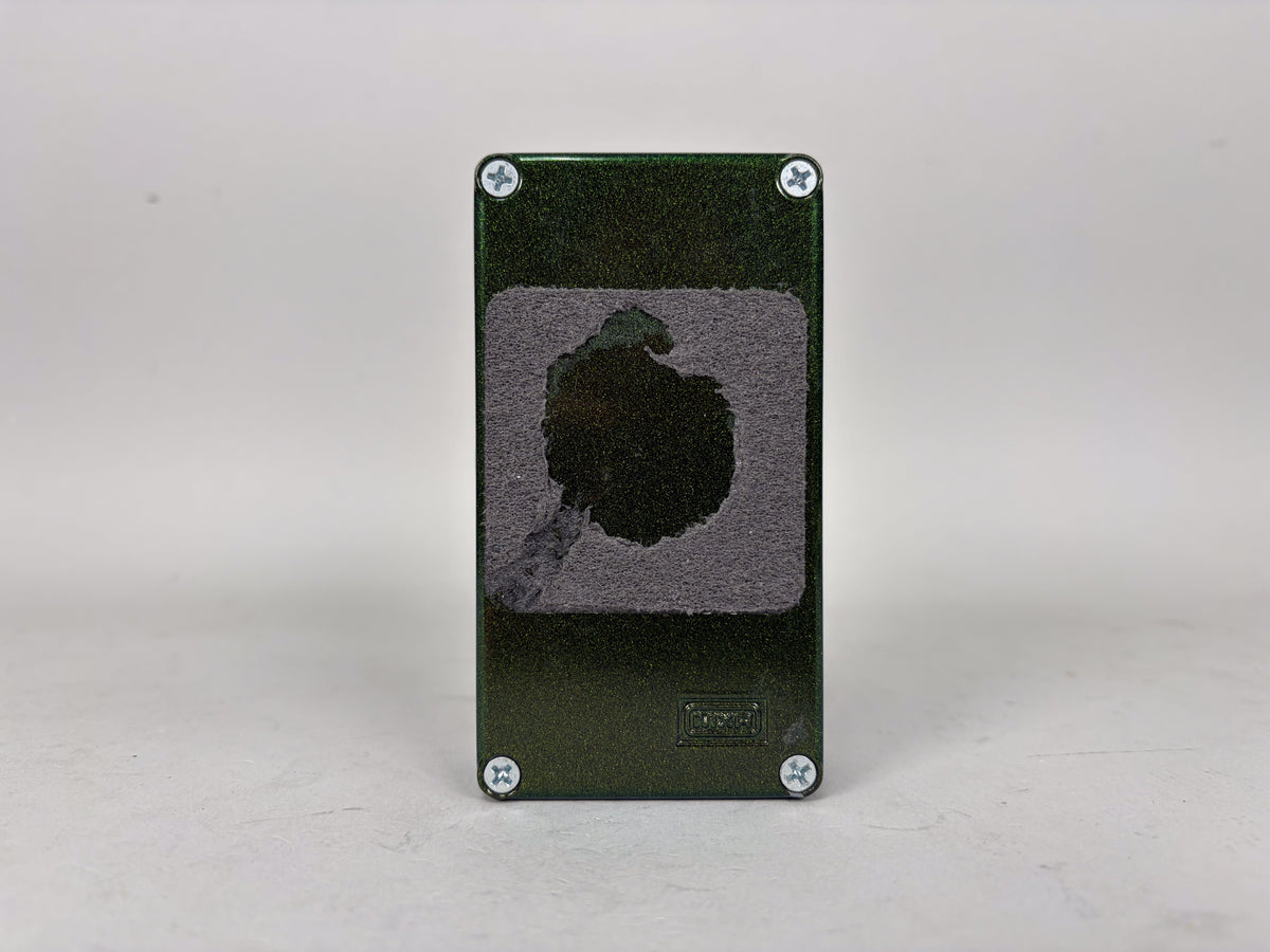 The dark green metallic pedal is viewed from the bottom, showing four silver screws and a large, worn grey velcro patch.