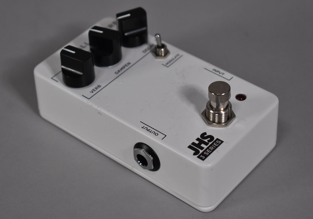 A white effects pedal, angled from the left, showing three black control knobs, a toggle switch, a footswitch, and an output jack.