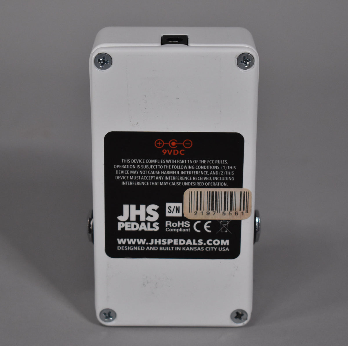 The white pedal is viewed from the back, showing a black label with white text and a barcode, secured by four silver screws.