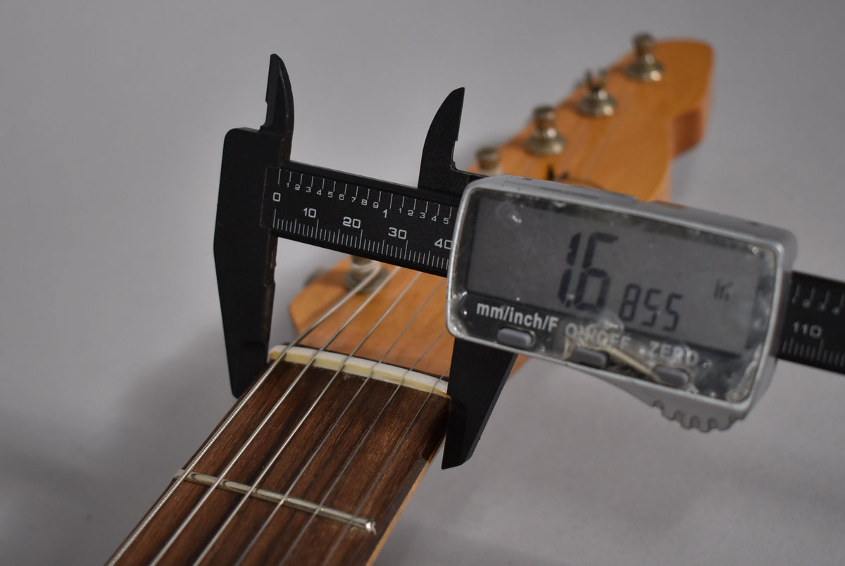 A digital caliper measures the guitar's nut width at 1.6855 inches, with the headstock and tuners blurred in the background.