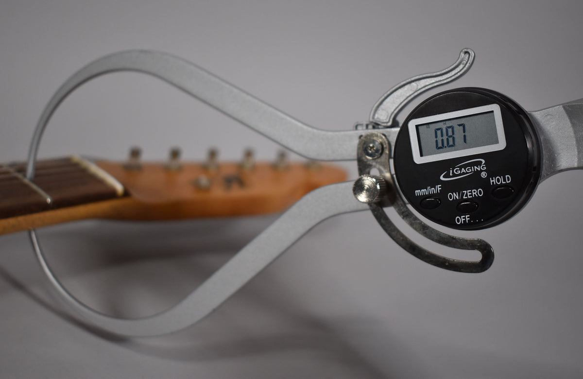 A digital caliper measures the guitar's neck, showing "0.87" on its screen, with the headstock and tuners blurred in the background.