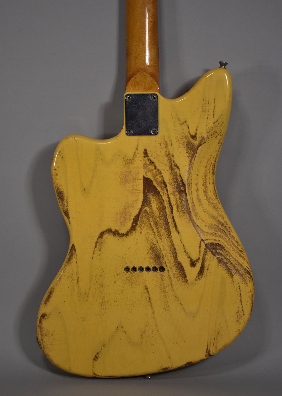 The back of a butterscotch blonde electric guitar with heavy brown relicing, a neck plate, and six string ferrules.
