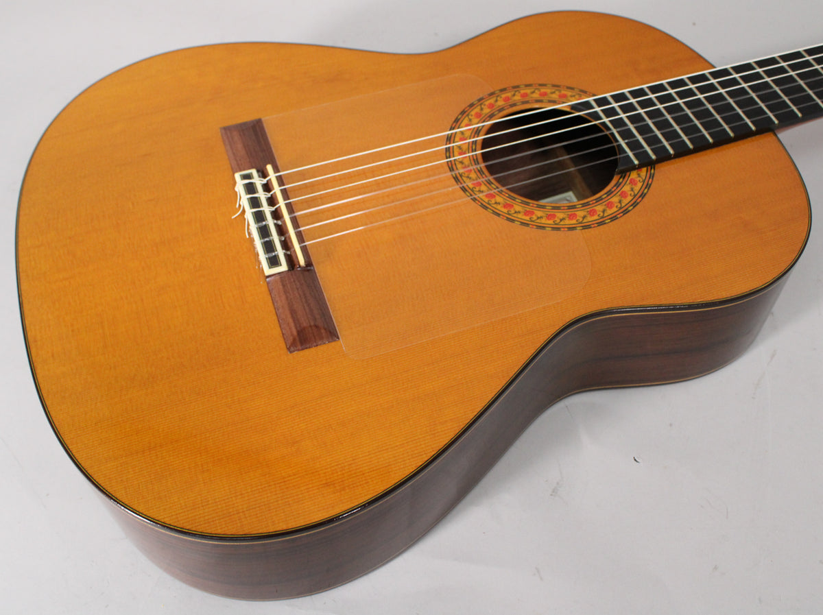 The classical guitar's light-colored top, dark sides, and ornate rosette are visible from an upper-right angle, with a clear pickguard.
