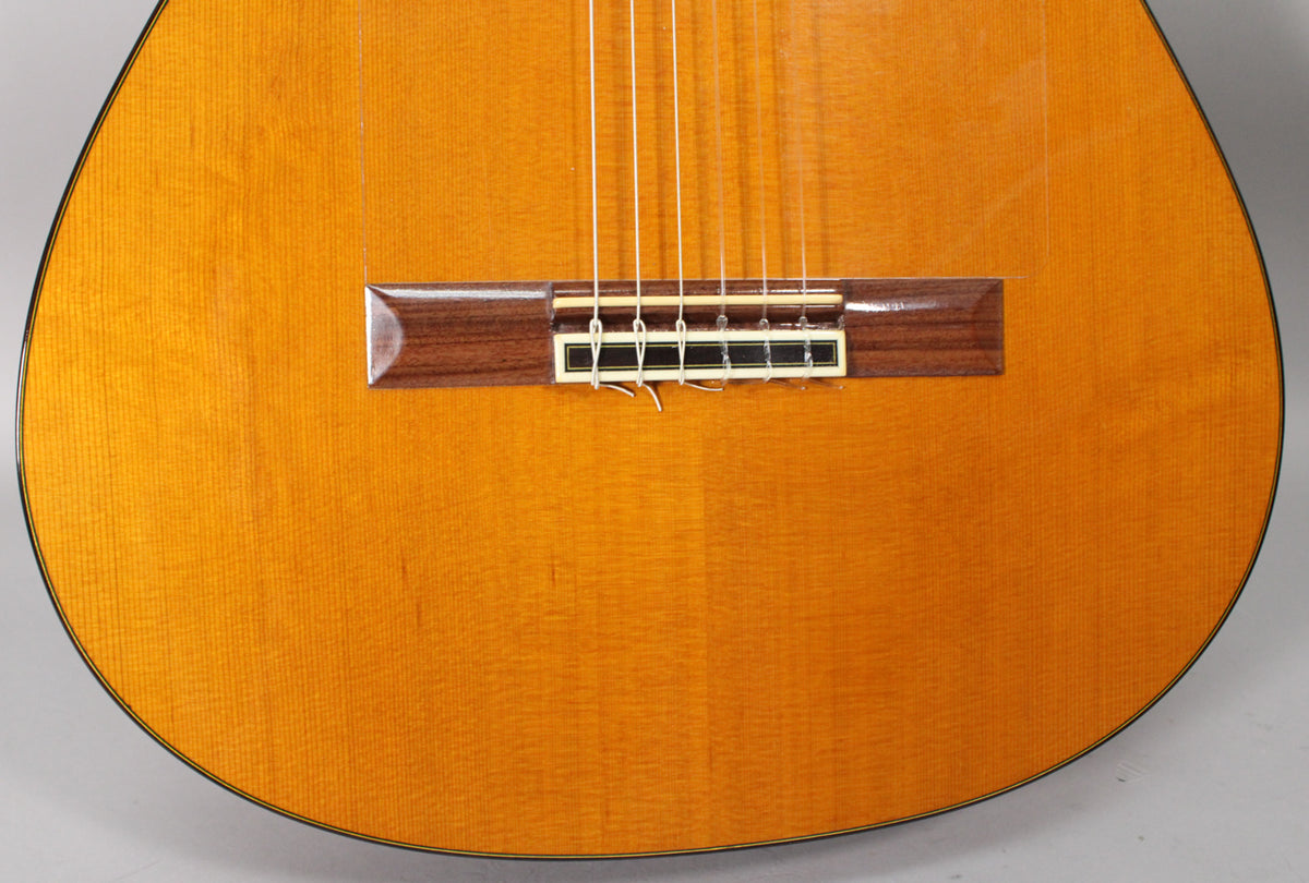 Close-up of the classical guitar's light-orange spruce top, showing the dark rosewood bridge with a white saddle and tied nylon strings.
