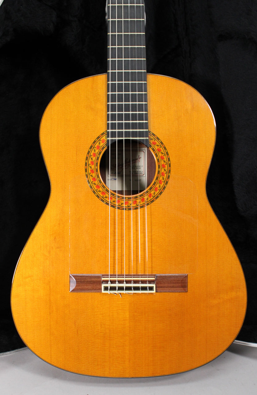 Full front view of the classical guitar, showing its light amber body, dark fretboard, and a colorful floral rosette around the soundhole.