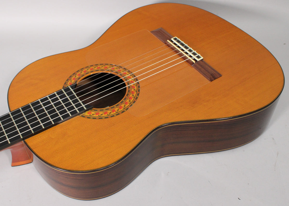 A classical guitar, angled from the upper left, shows its light brown top, dark wood sides, and a colorful rosette around the soundhole.