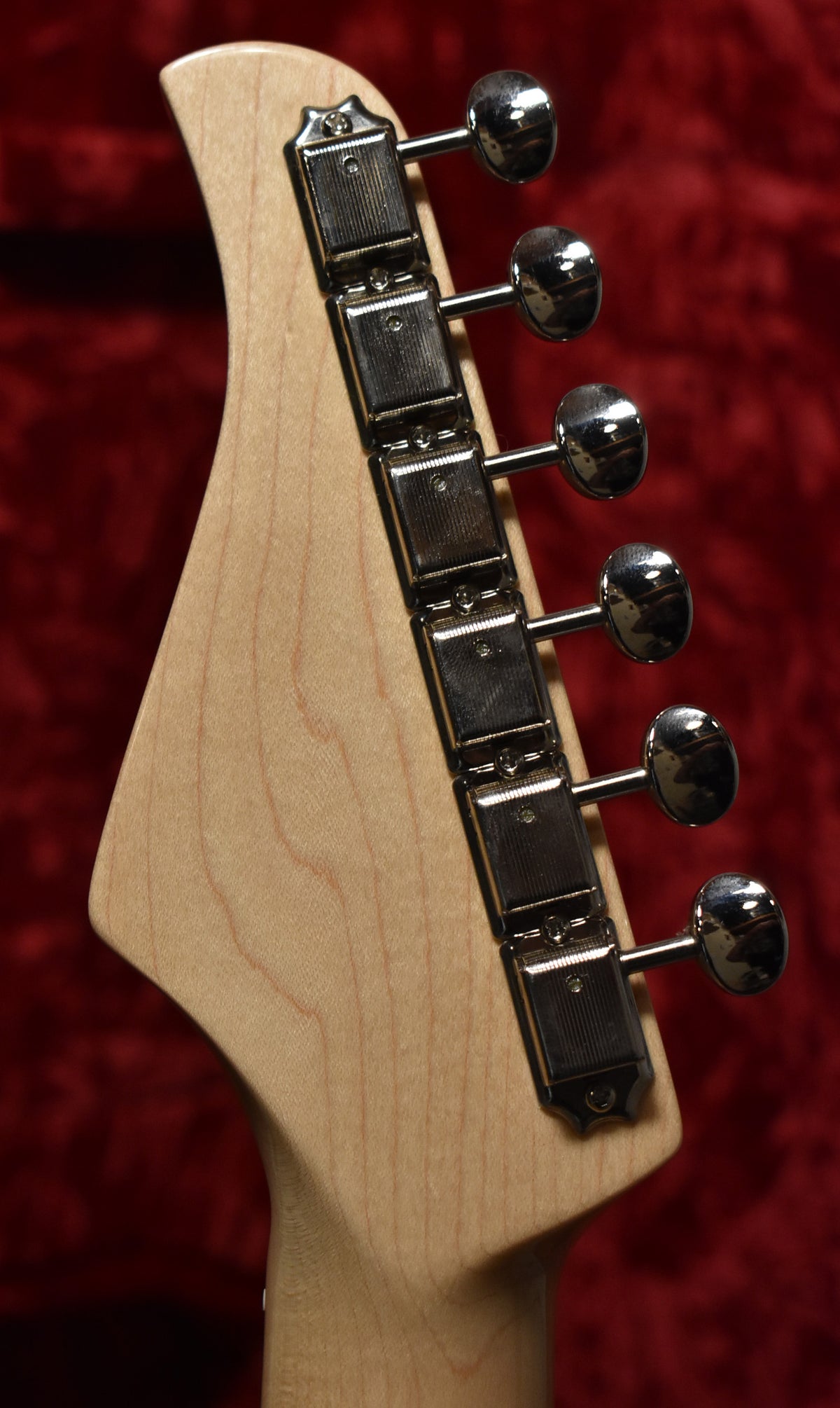 The guitar's light wood headstock, angled left, features six chrome tuners with oval buttons, against a red velvet background.