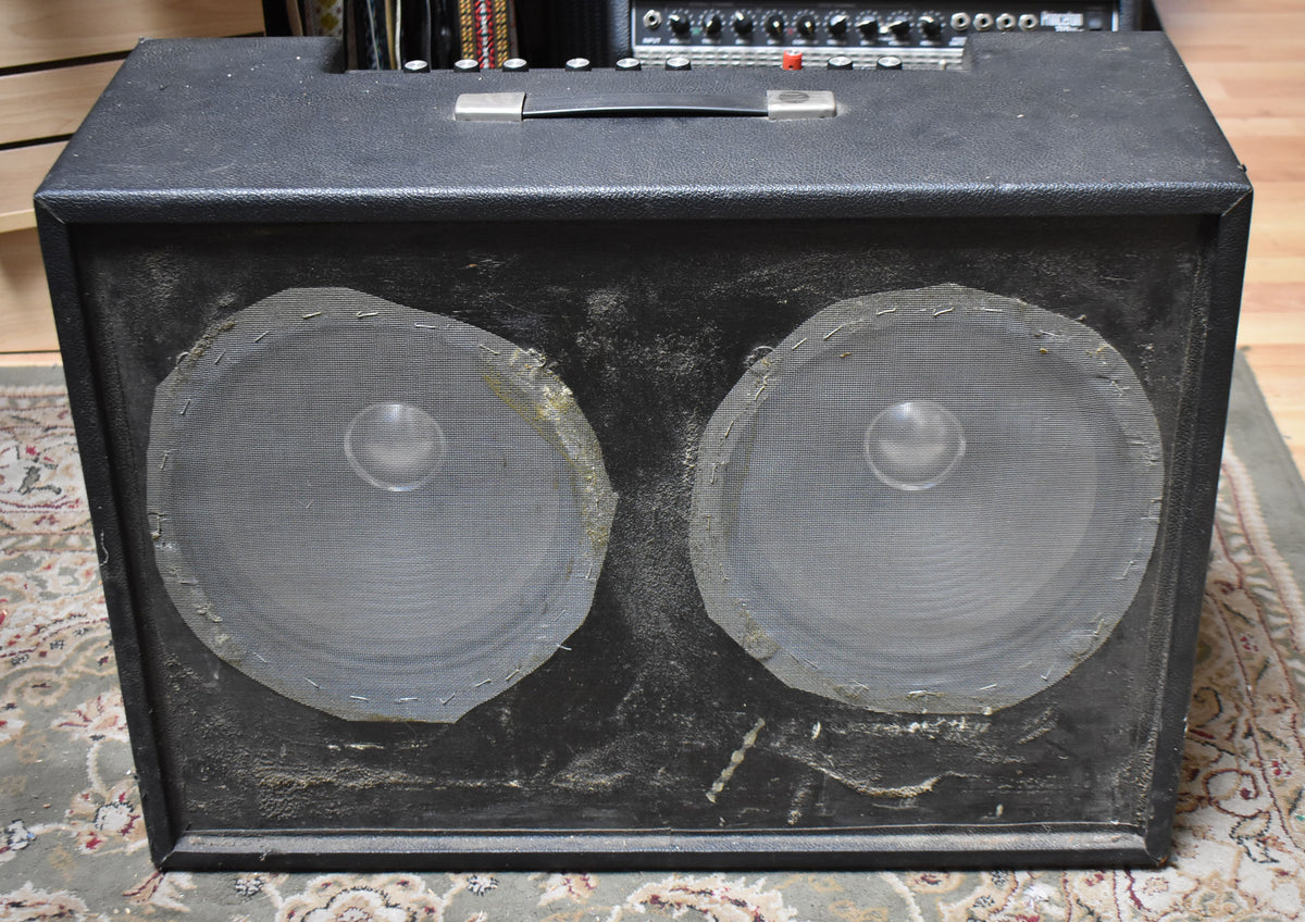 Front view of a black combo amp with two large, dusty speakers, their mesh grilles stapled to frayed baffles.