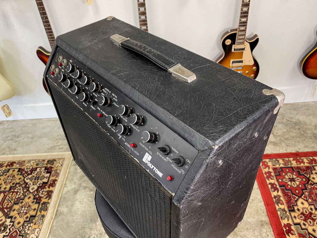 Angled view of a black combo amp with a textured finish, showing the top handle, metal corner protectors, and numerous black control knobs.