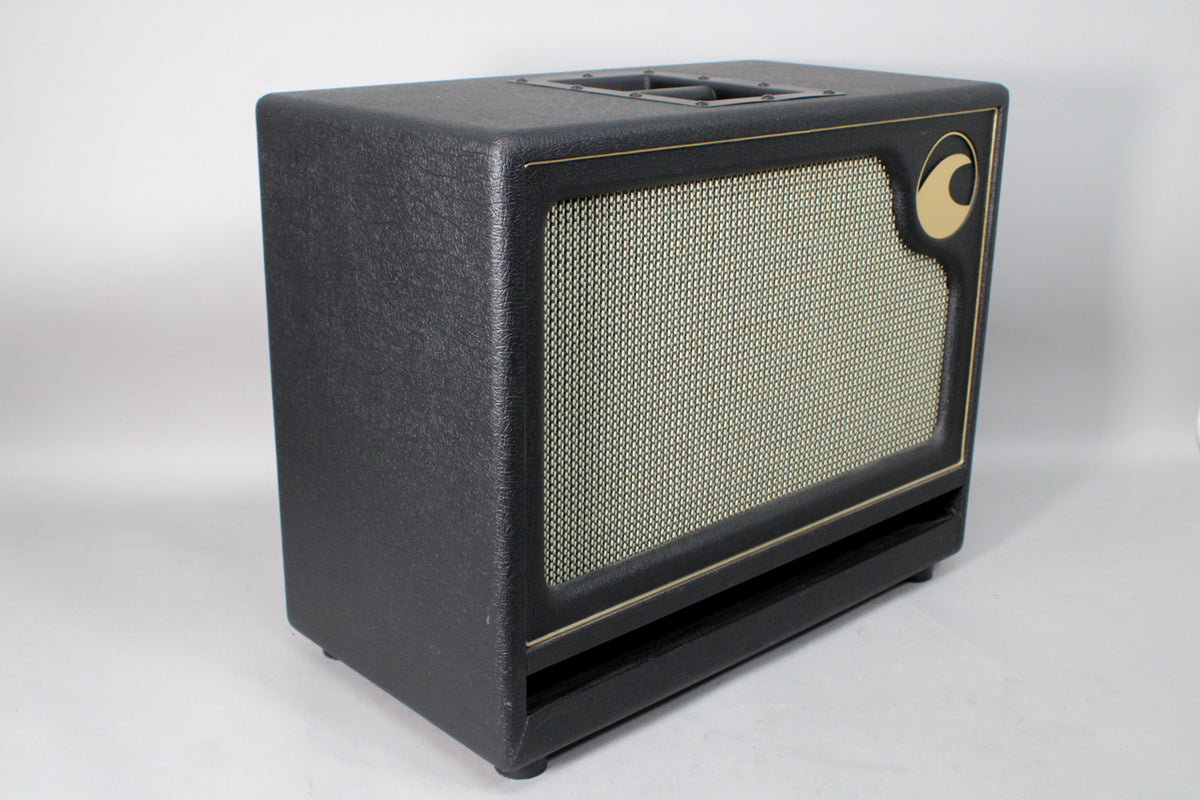 Black textured speaker cabinet, angled from the right, with a light tan grille, gold piping, and a gold crescent logo.
