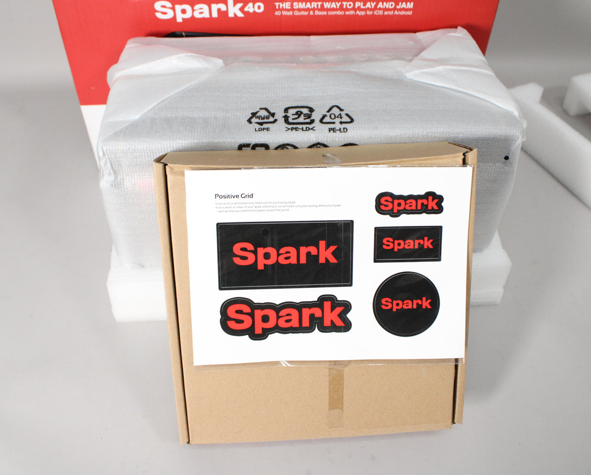 The amp is partially visible, still in its plastic wrap and protective foam, with a brown cardboard box and sticker sheet in the foreground.