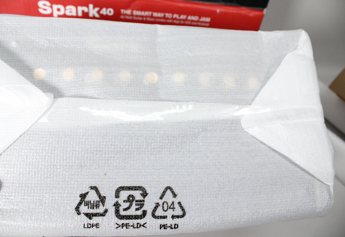 The amp is wrapped in white, textured plastic, with recycling symbols visible on the bottom edge, and a red box in the background.