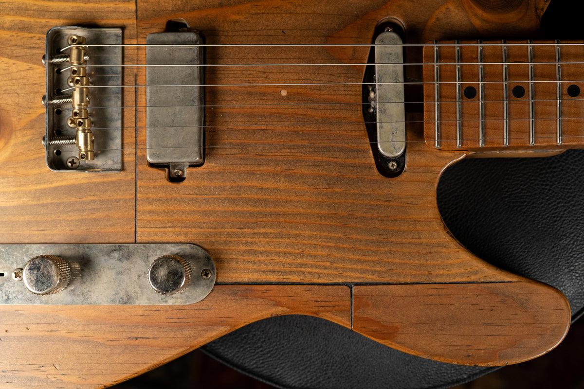 Close-up of a light brown pine guitar body with visible grain, showing the bridge, two pickups, and two worn metal control knobs.