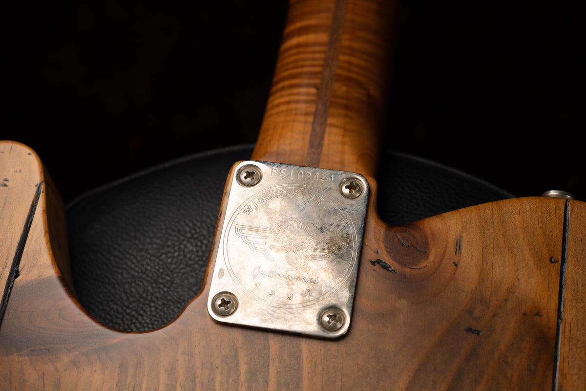 Close-up of the guitar's neck joint, showing a worn, engraved metal neck plate screwed into the dark, distressed pine body.