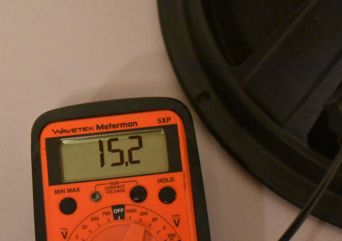 An orange multimeter displays "15.2" on its screen, with part of a black speaker frame visible in the upper right.