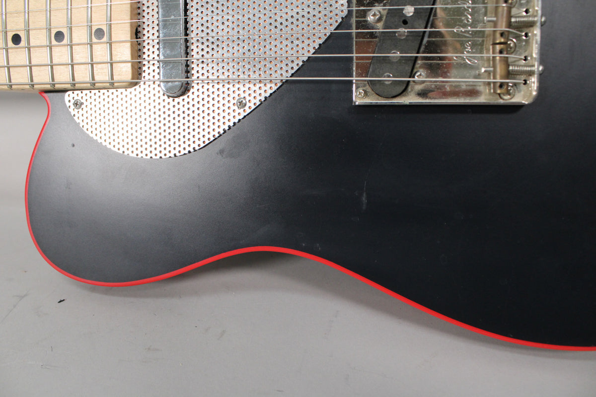 Close-up of the matte black body with red binding, showing a perforated metal pickguard and chrome bridge with three barrel saddles.