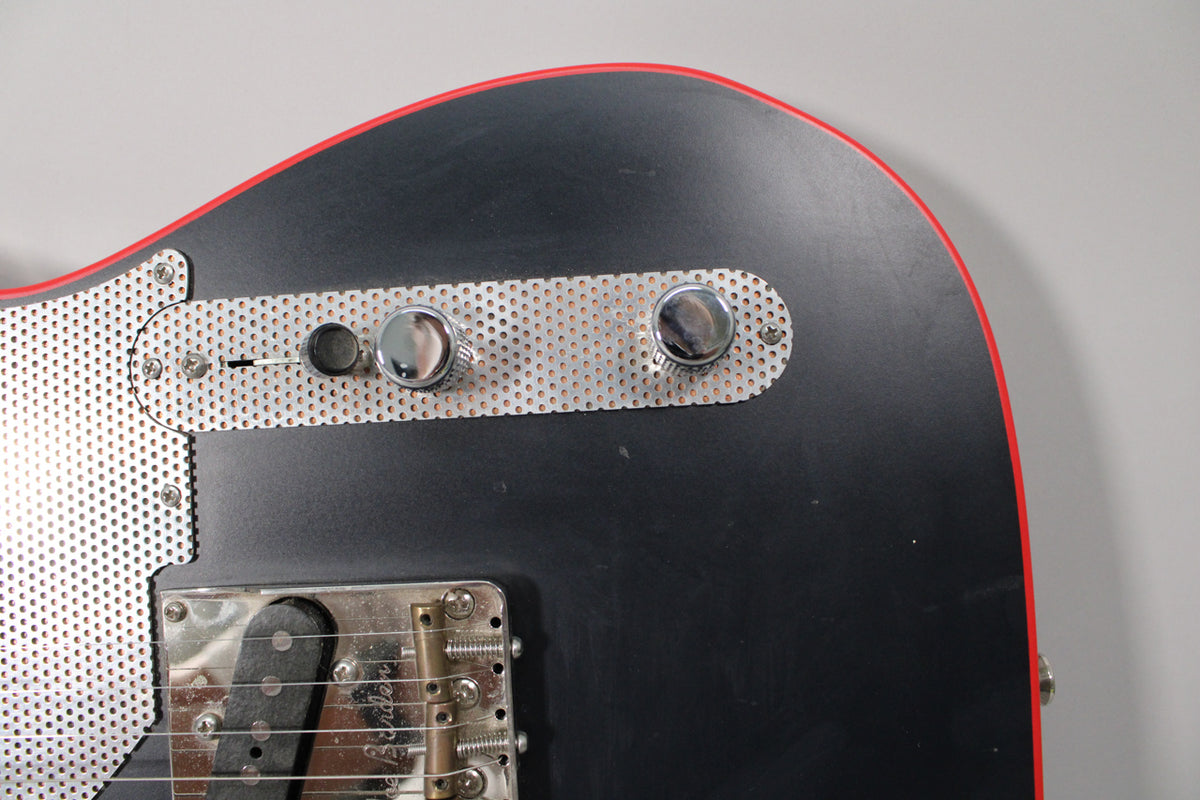 Close-up of the matte black guitar body with red binding, showing the perforated metal control plate and bridge.