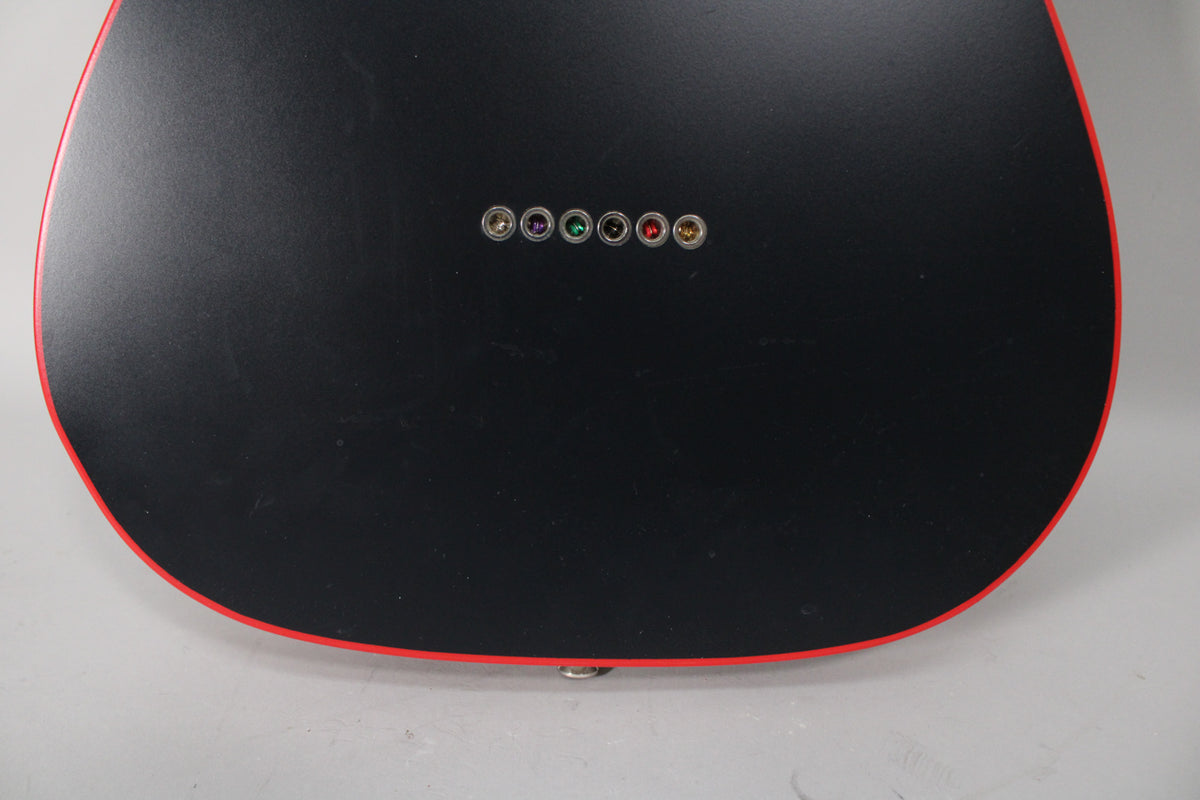 The matte black guitar back, framed by red binding, shows six string ferrules with colorful centers and light surface scuffs.
