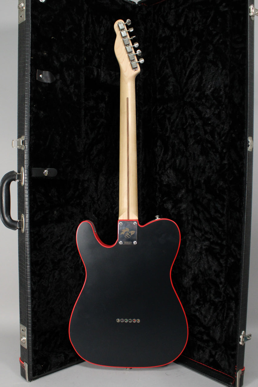 The guitar's matte black body with red binding is seen from the back, standing in its open, black-lined hard case.