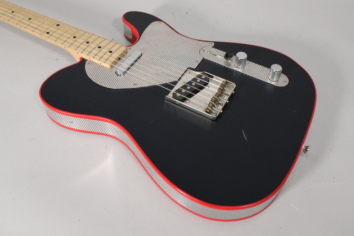 Angled view of a matte black guitar with red binding, perforated metal pickguard and side trim, and visible scratches on the body.