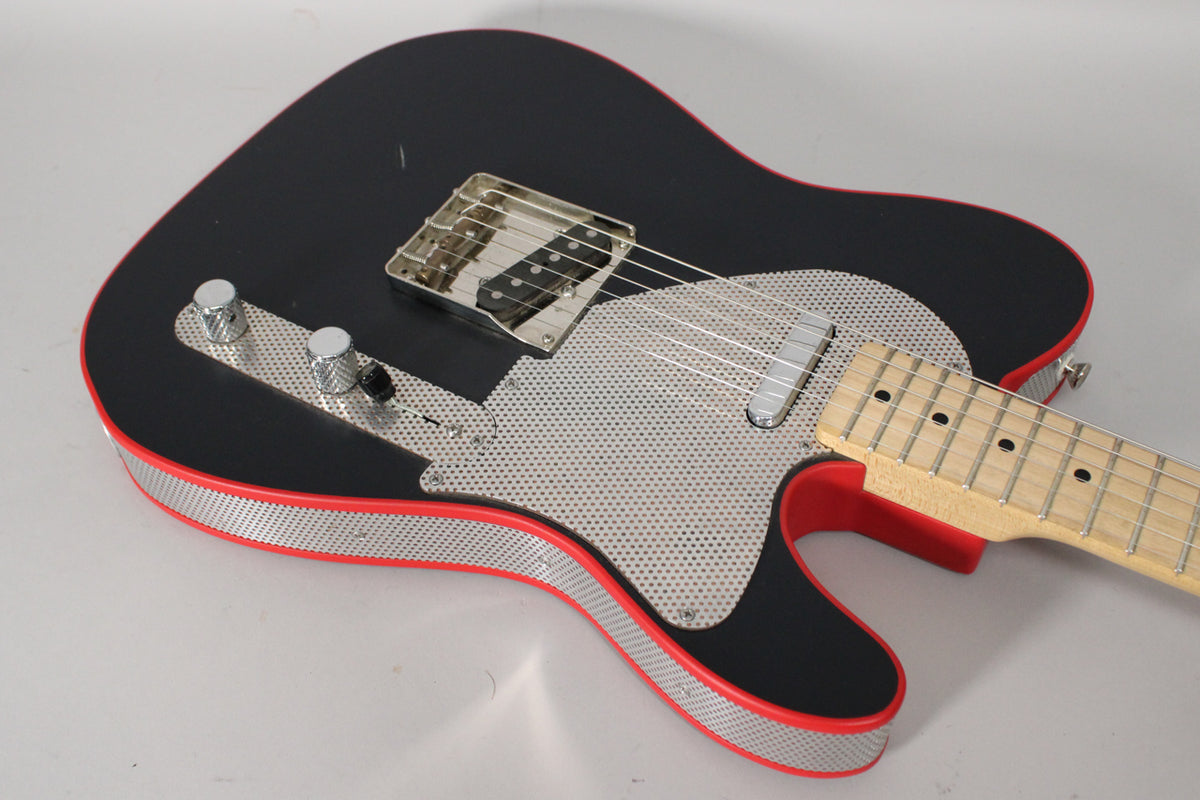 The matte black guitar body, with red edges and perforated metal sides, is angled to show the chrome bridge pickup and controls.