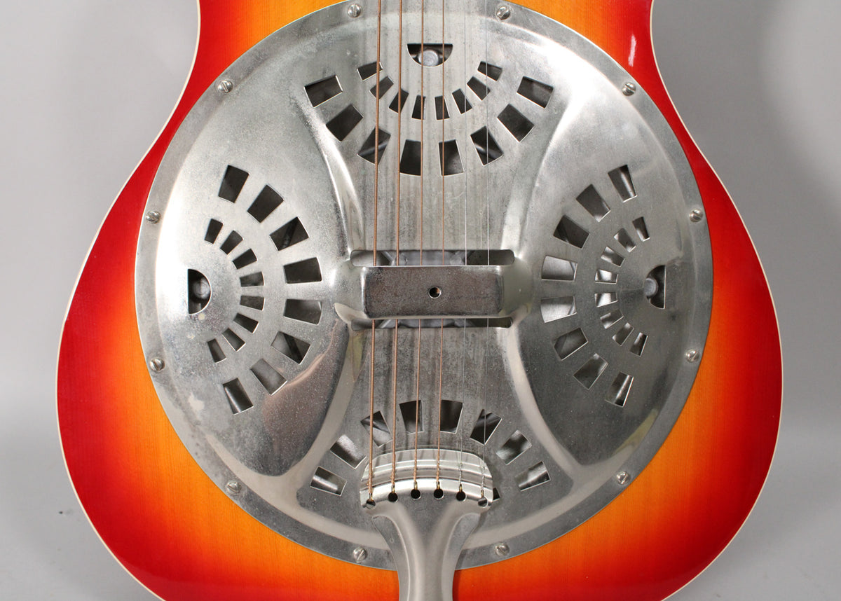 Close-up of a resonator guitar's cherry sunburst body and metal resonator cover with decorative cutouts, strings, and bridge.